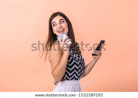 Indoor picture of young good-looking European woman isolated on peach background holding coffe to go and phone in hand and smiling while browsing or reading message