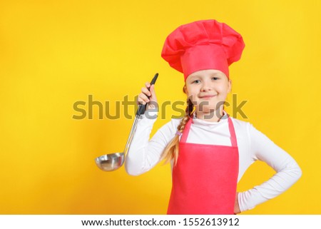Cheerful cute baby chef holds soup ladle in hands. Little girl in uniform on a yellow background.