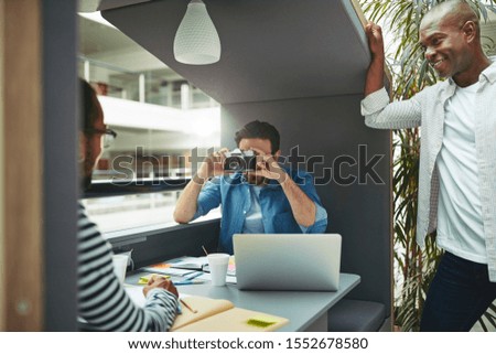 Designer taking a photo of a colleague with a vintage camera while working together in a meeting pod in a modern office