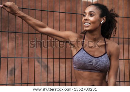 Attractive smiling healthy confident fit african sportswoman resting after workout outdoors at the sports ground, listening to music with wireless earphones