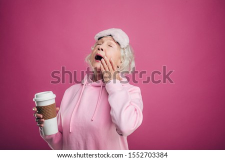 Picture of sleepy grandma with sleeping mask on forehead and cup of drink in hand. Yawn. Wear pink modern sweater. Isolated over pink background