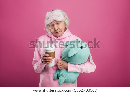 Confident but weird old senior woman stand alone and show cup or thermos in hand. Embrace green toy with another hand. Sleeping mask on forehead. Wear hoody or sweater. Isolated on pink background