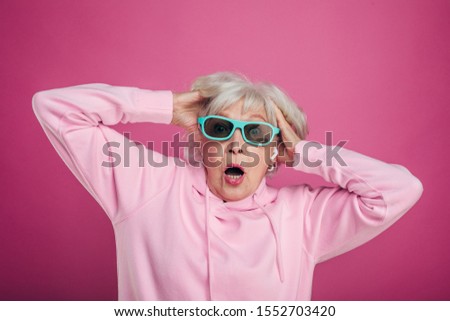 Terrified and scared old woman hold hands in hair and look straight through sunglasses. Wear pink hoody. Emotional stressed. Isolated over pink background