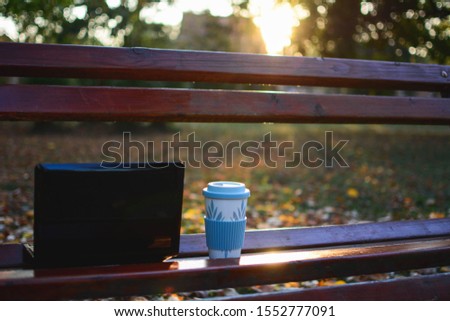 Laptop and a cup of coffee on a wooden bench in the park before sunset in the fall.