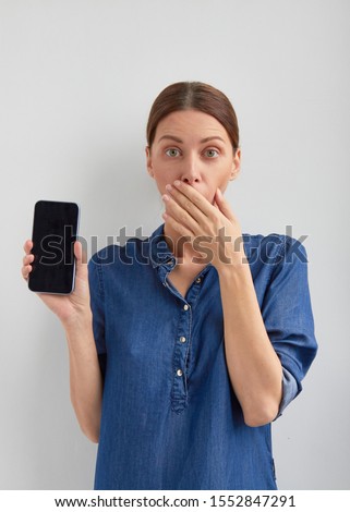 Shocked startled young brown tucked back hair girl in jeans holds in hand phone and shows smartphone black empty not working screen and closing surprised opened mouth with big opened eyes Royalty-Free Stock Photo #1552847291