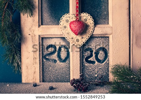 Christmas New Year decor - frame of snow-covered window with hand drawn 2020, fir branches, cones, and two decorative hearts. Cottage interior. Royalty-Free Stock Photo #1552849253