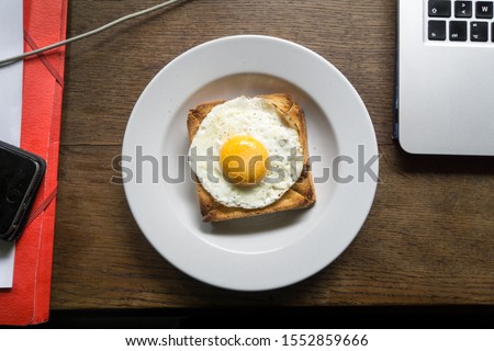 High angle close up view over a toasted cheese with ham sandwich and a fried egg on the top, in a white plate on a wooden desk with cable, file and devices around, lunch time at work in France.