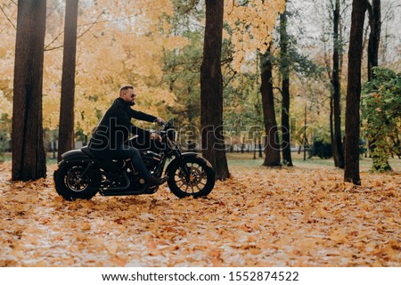 Handsome carefree bearded motorcyclist rides fast motorbike, wears shades, rides bike in park, poses against yellow trees and foliage, empty space for your information. Motor tourist outdoor