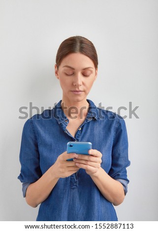 Isolated young beautiful woman with serious face in blue jeans dress holds in hands smartphone and uses phone by fingers to type answer or order action in application with joy on white background Royalty-Free Stock Photo #1552887113