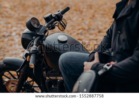 Cropped shot of faceless biker poses on fast motorbike, stops in park, ground covered with fallen leaves, enjoys travel and journey. People, lifestyle, transport concept.