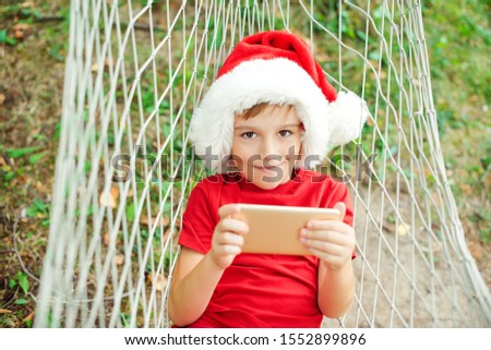 ?ute little boy watching christmas cartoons on family vacations. Happy kid in Santa hat lying in hammock. Family travelling to tropical countries. Happy child using mobile phone