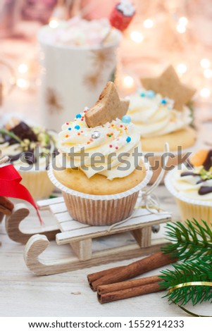 Christmas decorated cupcakes on wooden sledge. Cinnamon rolls, cotton flowers, tangerine bites, christmas tree branches and gingerbread cookies. String lights and cup with marshmallow on background.