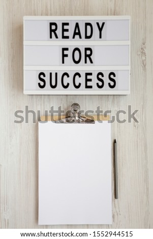 'Ready for success' words on a lightbox, clipboard with blank sheet of paper on a white wooden background, top view. Overhead, from above, flat lay. 