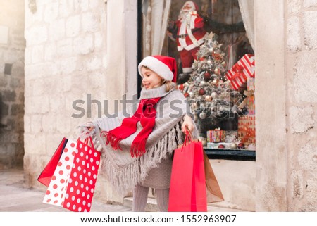 Happy child is enjoying Christmas shopping and gifts. Kid girl with red paper bags is buying presents on market street by store showcase in Provance. Cozy fair, New Year and Black Friday