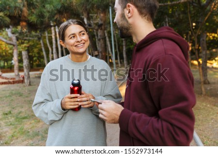 Image of a young pleased cheery happy positive sports woman and man using mobile phone outdoors in nature green park holding bottle with water.