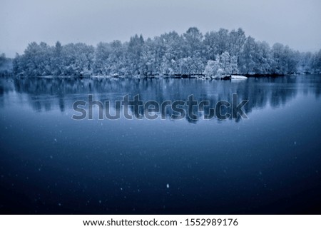 Snow falling on deep blue cold water in front of beautiful snow covered trees and landscape.
