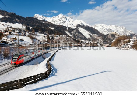 A local train traveling by Salzach River through the snowy valley on a sunny winter day, with Hohenwerfen Castle perched on a hilltop under alpine mountains in background, in Werfen, Salzburg, Austria