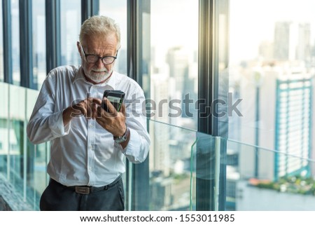 Picture of old business man with a phone in his hand. He is in his 70th with grey hair. He is wearing a white shirt and black trouser. He stands by the window with city view outside.