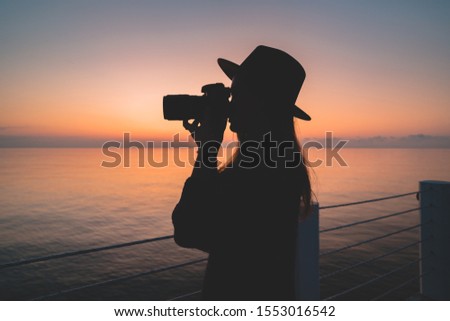 Silhouette of a woman photographer in hat with dslr camera during taking pictures of sea at sunset