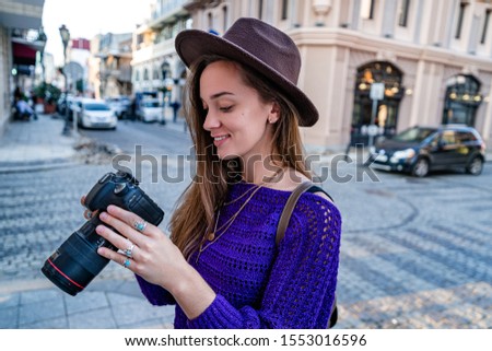 Happy beautiful casual stylish hipster traveler woman photographer in hat looking photos on dslr camera after taking pictures while walking around the european city
