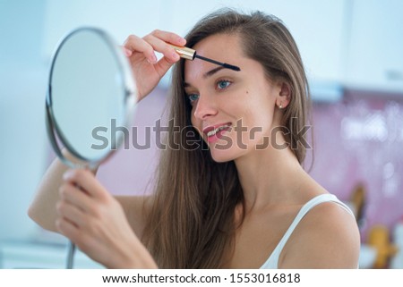 Beautiful attractive smiling brunette woman uses a small round mirror and puts black mascara on her eyelashes during home makeup in the morning