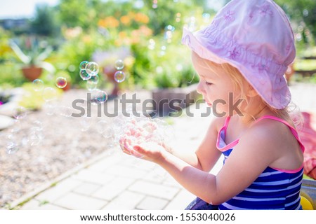 Adorable blonde baby girl 3 year old in a pink hat and blue stripped swimsuit having bath at backyard and playing with bubbles.
