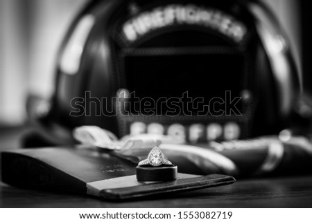 A teardrop, or pear, shaped diamond engagement ring on top of a wedding band and fireman's ax with a firefighter helmet blurred in the background. Black and white.