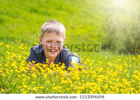 A boy with a smile is resting on a green lawn with yellow wildflowers in the sunshine. Smiling little boy lying on the grass with flowers in the park, springtime