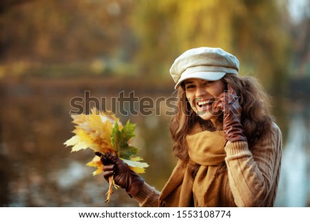Hello autumn. Portrait of smiling trendy middle age woman in sweater, hat, gloves and scarf with yellow leaves speaking on a smartphone while sitting on a bench outdoors in the autumn park.