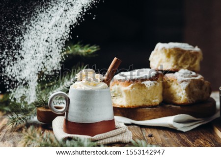 A hot cocoa with whipped cream, cinnamon stick and fresh Christmas buns with powder and over festive table, sugar powder is sprinkled on the background lke a snow. Winter holidays.