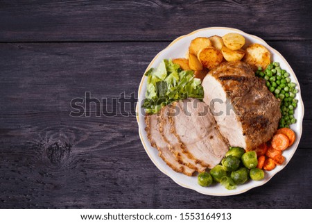 Roasted sliced ham and vegetables: potatoes, carrots, brussel sprouts, cabbage and green peas on black wooden background. copy space