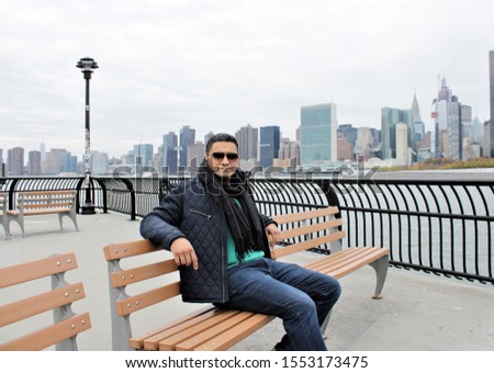A handsome Indian man sitting on a bench on a waterfront in front of Manhattan Skyline.