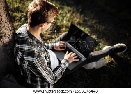 Back view photo of handsome young male student in casual outfit using notebook or laptop while sitting on the grass at the park.