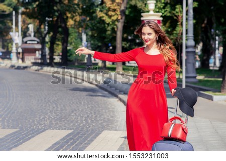 Tourist business woman in beautiful red long dress with suitcase is catching taxi on the city street.