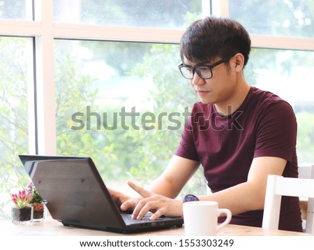 Asian young man wearing eye glasses sitting in the coffee shop looking  thoughtfully at screen of  computer laptop on the table ,  Royalty-Free Stock Photo #1553303249