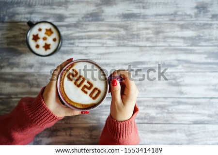 Coffee cup with number 2020 on frothy surface in female hands holding over grey painted wood plank background and another cup with star symbols on frothy surface. Happy new year 2020 food art theme. Royalty-Free Stock Photo #1553414189