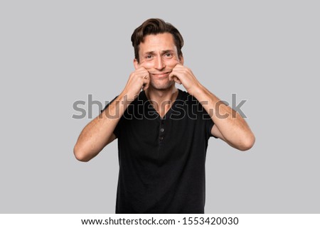 Man in a black tee shirt stretching lips into a an artificial smile. Being incencere. 