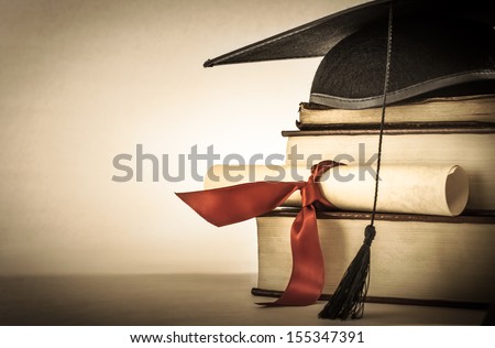 A mortarboard and graduation scroll, tied with red ribbon, on a stack of old battered book with empty space to the left.  Slightly undersaturated with vignette for vintage effect. Royalty-Free Stock Photo #155347391
