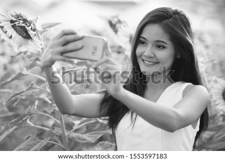 Young happy Asian woman smiling while taking selfie picture with mobile phone in the field of blooming sunflowers