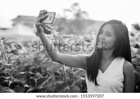 Young happy Asian woman smiling while taking selfie picture with mobile phone in the field of blooming sunflowers