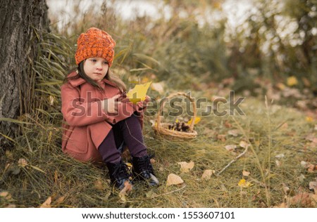 sad preschooler girl in an orange hat and a brown coat sits on the ground under an old big tree. autumn horizontal photo of a child.