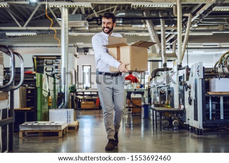 Smiling caucasian bearded graphic engineer in shirt and tie walking in printing shop and relocating box. In background are printing machines. Royalty-Free Stock Photo #1553692460