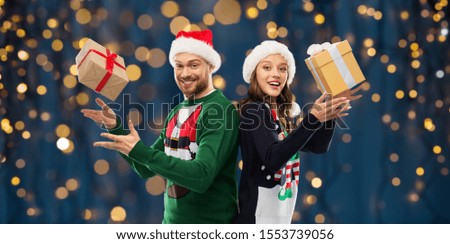 people and holidays concept - happy couple in santa hats with christmas gifts at ugly sweater party over festive lights on dark night background