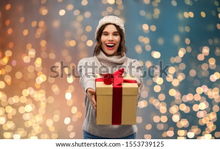 christmas, holidays and people concept - happy smiling young woman in knitted winter hat and sweater holding gift box over festive lights background