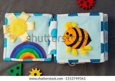 Handmade textile book for baby on gray background. Pages with funny felt bee, woolly rainbow and sun. Baby development. Educational toys. Fine motor skills.