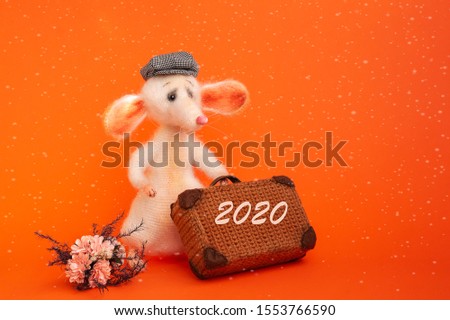 White, knitted mouse with a suitcase in his paw, in a plaid cap. A bouquet of flowers lies on the side of the figure. Symbol of a new two thousand twentieth year. Isolated bright orange background.