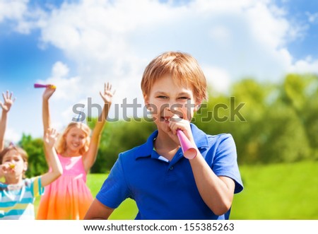One happy boy blows noisemaker horn on a birthday party wearing cap with friends standing on background