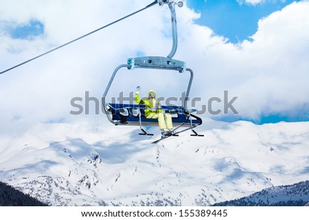 Happy skier young woman sit on the ski lift and waiving hand with mountains on background