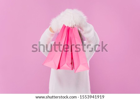 Girl holding pink paper bags over isolated pink background. A girl holding packets at the head covering his face. A girl in a sweater and fluffy mittens.