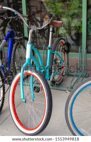 A trendy teal-colored bicycle is chained up among other bikes on a rack outside of a building. It has red and white lining on its tires and a vintage look.